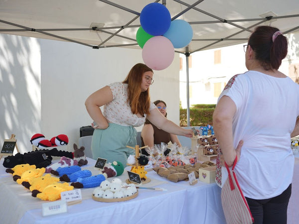 Two women interacting at a table with crochet plushies and various items under a tent with colorful balloons.